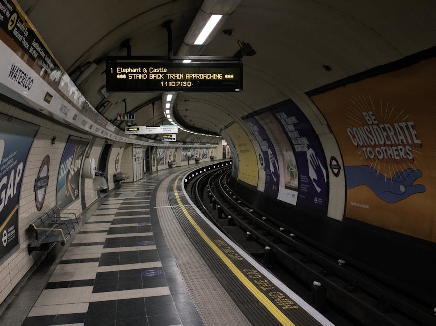 Interior view of an underground train station platform at Elephant and Castle, featuring an arched ceiling with fluorescent lighting. The platform has a black and white tiled floor, and there are two empty benches along the wall. An electronic sign displaying the message 'Elephant & Castle *** STAND BACK TRAIN APPROACHING ***' is suspended above the platform, showing the current time as 11:07:30. The curved train tracks extend into the tunnel, with curved yellow and white safety markings along the platform edge. On the wall to the right, there are large colorful posters, including one with the message 'BE CONSIDERATE TO OTHERS' accompanied by an illustration of a hand. This environment is typically associated with transport and logistics, reflecting a setting where items or furniture might be transported during home relocation or moving services provided by companies like manwithavanelephantandcastle.co.uk, especially when coordinating the loading and transportation of household belongings.