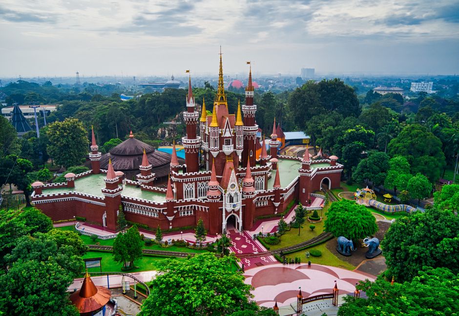 An aerial view of a colorful fairy tale castle situated within a theme park, surrounded by lush green trees and well-maintained landscaping. The castle features multiple tall spires with orange and yellow roofs, white decorative accents, and a central entrance with a pointed archway. In the background, there are dense wooded areas and distant cityscape elements under a partly cloudy sky. The park appears to have various walkways, small structures, and attractions at its perimeter, with visible pathways leading towards the castle. This setting captures the intricate details of the castle's architecture and the vibrant environment of the theme park, reflecting a place designed for entertainment and family visits. The image suggests a scenic and lively location, ideal for family trips or leisure activities, with the castle as the focal point of the attraction and surrounded by themed playground features and trees.