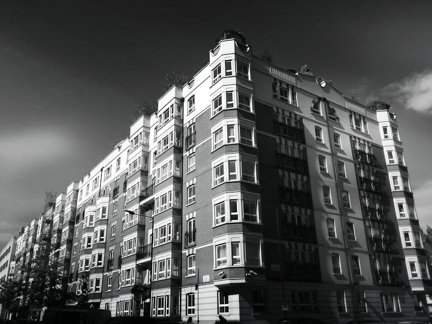 Black and white photograph of a large multi-storey residential building located in an urban area, with numerous windows and balconies visible on all floors. The building has a modern architectural sty