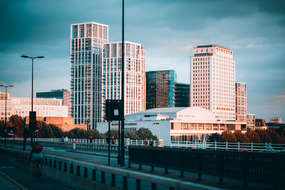 A view of modern high-rise buildings with glass facades and white concrete frames, situated in an urban cityscape during late afternoon or early evening, with the sky showing a mix of clouds and soft lighting. In the foreground, a cyclist is riding on a multi-lane road separated by white and black striped barriers, with traffic cones along the side. There is a white fence lining the sidewalk, and several pedestrians are visible walking near the buildings. The scene includes street lamps and trees along the lower part of the image. This photograph depicts an urban area suitable for house removals and furniture transport, highlighting the city environment relevant to home relocation services such as those offered by manwithavanelephantandcastle.co.uk, with visible infrastructure for moving logistics.