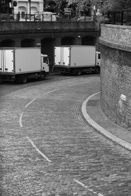 A black-and-white photograph showing a curved cobbled driveway or loading area adjacent to a brick wall and railing, with two white box trucks parked side by side in the background. The trucks are positioned for loading or unloading, with the rear doors closed. Inside the vicinity, there are no visible furniture or boxes, but the scene suggests a setting prepared for home relocation or furniture transport. The environment appears to be part of an urban area near residential or commercial buildings, with an overhanging balcony or terrace visible above the brick wall. The overall scene depicts the typical logistical arrangement involved in house removals or moving services, with trucks ready to carry household items via the driveway, which could be accessed for a home move. The area is well-lit, likely during daytime, and the scene reflects the vehicle parking and loading process common in professional removals.