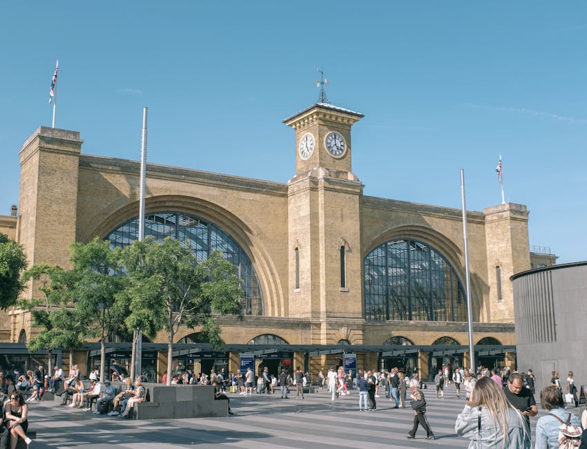 A large historic train station building with a beige brick façade, featuring two prominent arched glass windows and a central clock tower with a round clock face, located in a busy urban area. The entrance area is filled with pedestrians, some sitting on benches and others walking, with a row of ticket counters visible beneath the arched windows. In the foreground, there are several flagpoles without flags, a few trees providing some greenery, and a clear blue sky above. The scene depicts a bustling environment suitable for home relocation or furniture transport services, with the station serving as a reference point near Elephant and Castle, ready for loading or unloading of moving boxes and furniture as part of a professional removals process.